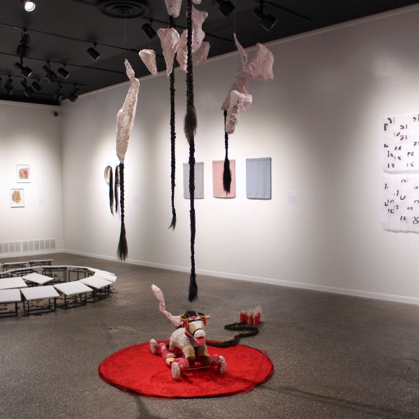 Gallery installation featuring suspended braided sculptures, textile and mixed-media wall works, a circular floor arrangement of small platforms, and a toy horse sculpture displayed on a red rug.