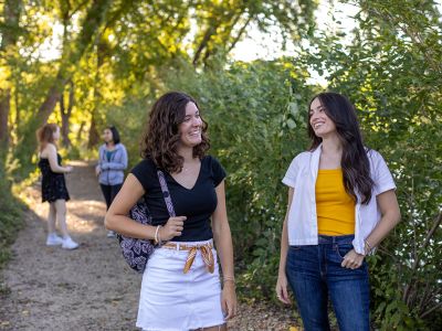 Four students walking on a wooded trail by a lake, talking and smiling.