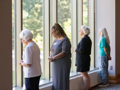 Four women stand looking out tall windows. 