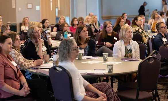 A group of people sit around tables, listening to a speaker (not pictured).