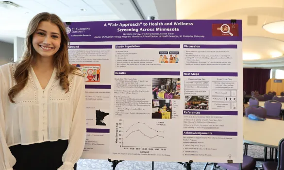 A student with long blonde hair smiles and stands in front of a poster board with research.