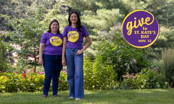 Two people wearing purple T-shirts and jeans stand in a green, leafy setting. A logo reads "Give to St. Kate's Day Nov. 12"