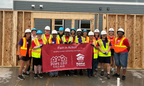 A group of people wearing neon safety vests and hard hats pose with a red banner at a construction site.