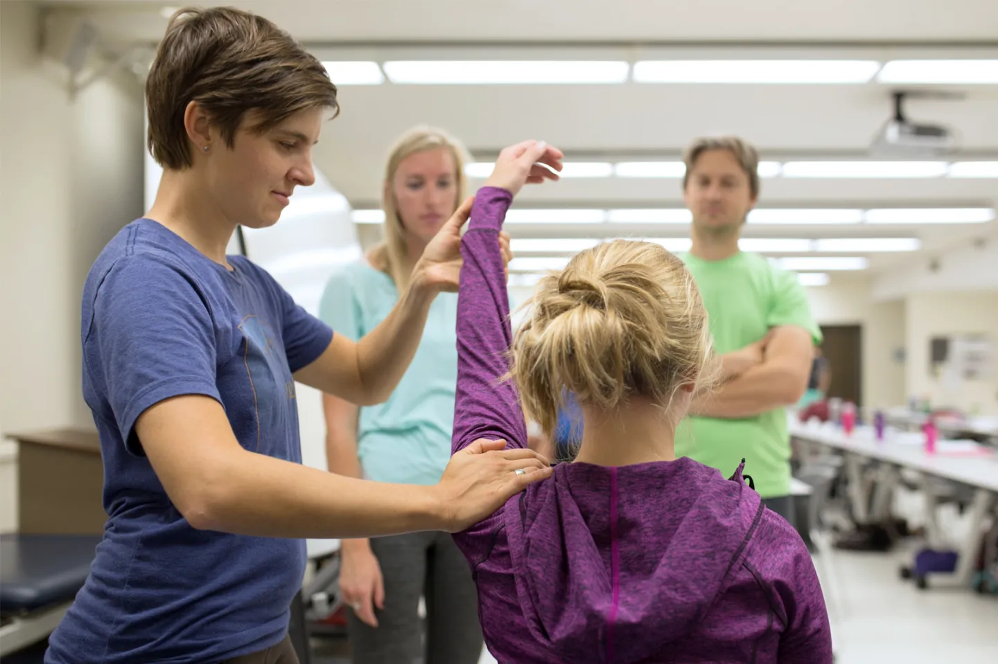 Doctorate of Physical Therapy student, wearing a blue shirt and standing, performing a health assessment on a seated patient in a purple long sleeve shirt. In a healthcare facility with two onlookers.