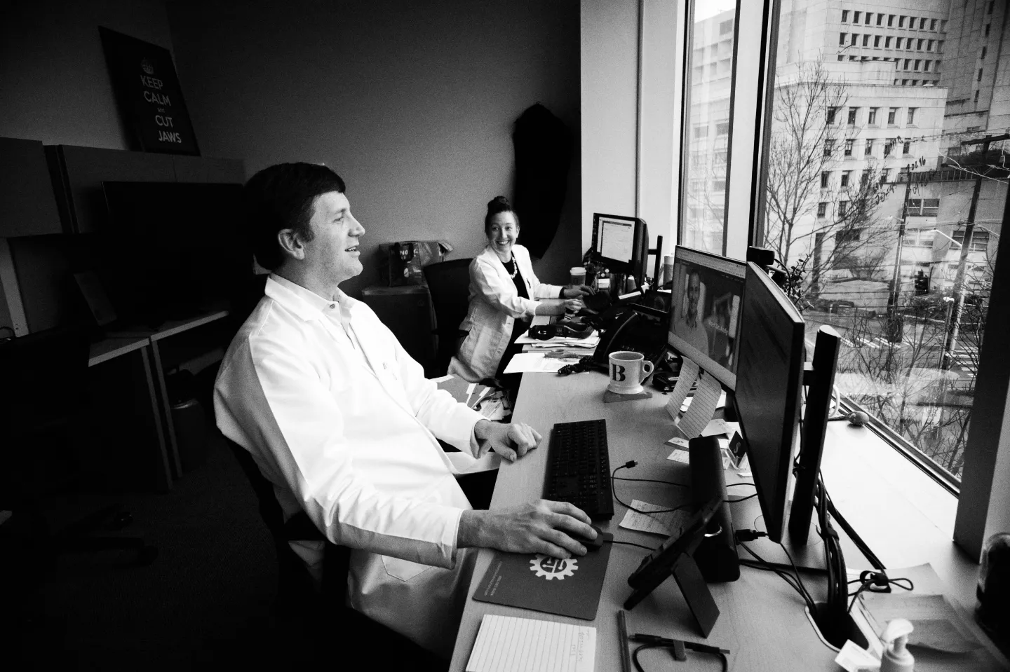 Black and white photograph of St. Catherine University alumna Andrea Burgess MPAS’15, PA-C, (right) and Dr. Sam Bobek, DMD, MD, FACS, in the Seattle Jaw Surgery clinic in Seattle, Washington. They are both smiling, seated in front of computers, facing a window. Photo Credit: Panravee Photography