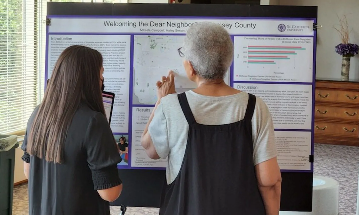 Two people, with their backs to the camera, look closely at a poster that reads "Welcoming the Dear Neighbor? Ramsey County"