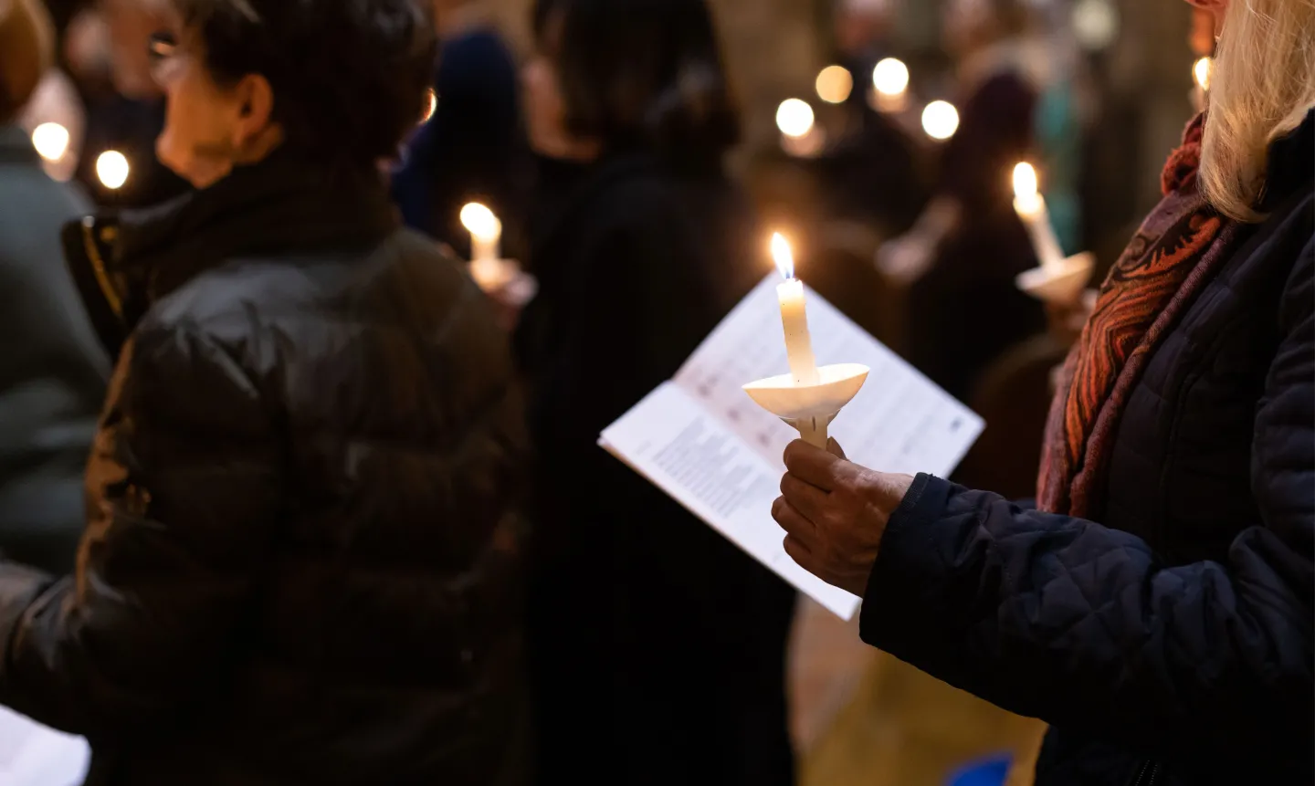A close-up picture of someone holding a lit candle and liturgical book. In the background, there are more lit candles and people.