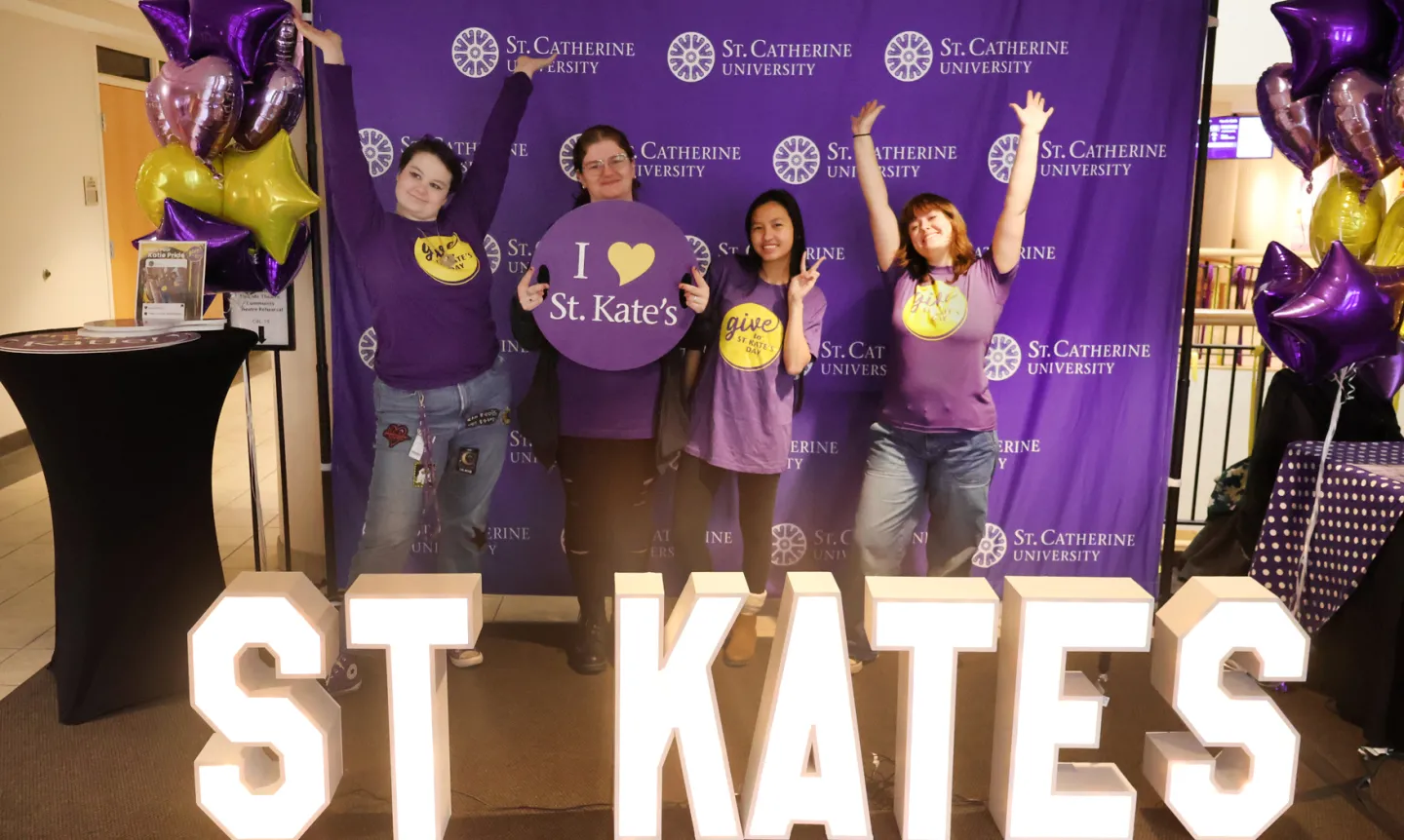 Four students in purple T-shirts stand behind glowing white letters that read "ST. KATE'S"