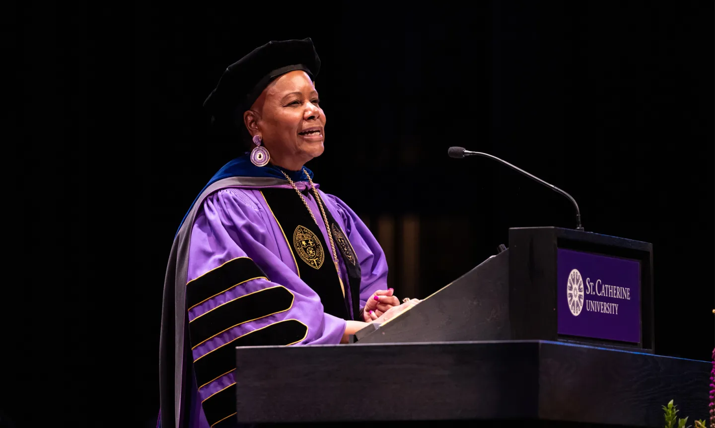 President Evans, wearing purple academic robes, stands before a microphone at a podium.