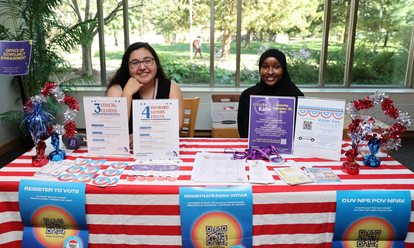 Two students sit behind a table with a red and white striped tablecloth and flyers about voting.