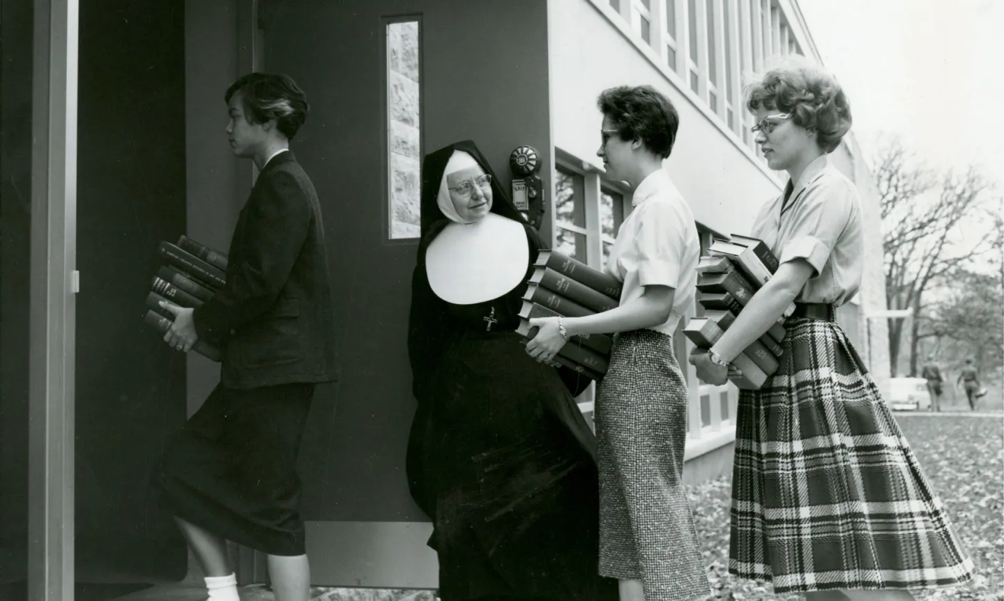 A black and white photo of a nun and three students carrying books into a building.