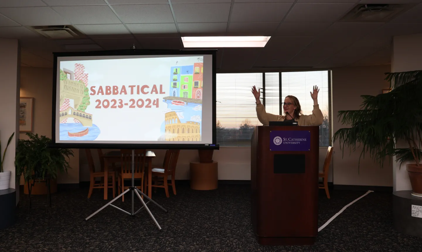 A screen features a slide that reads "Sabbatical 2023-24" and a woman behind a podium smiles and gestures.