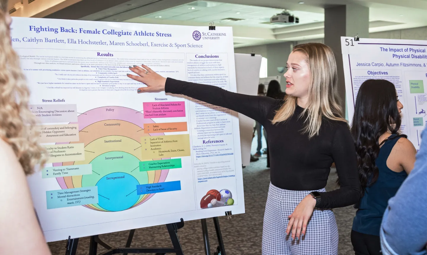 A student points at a research poster board with a multi-colored graph.