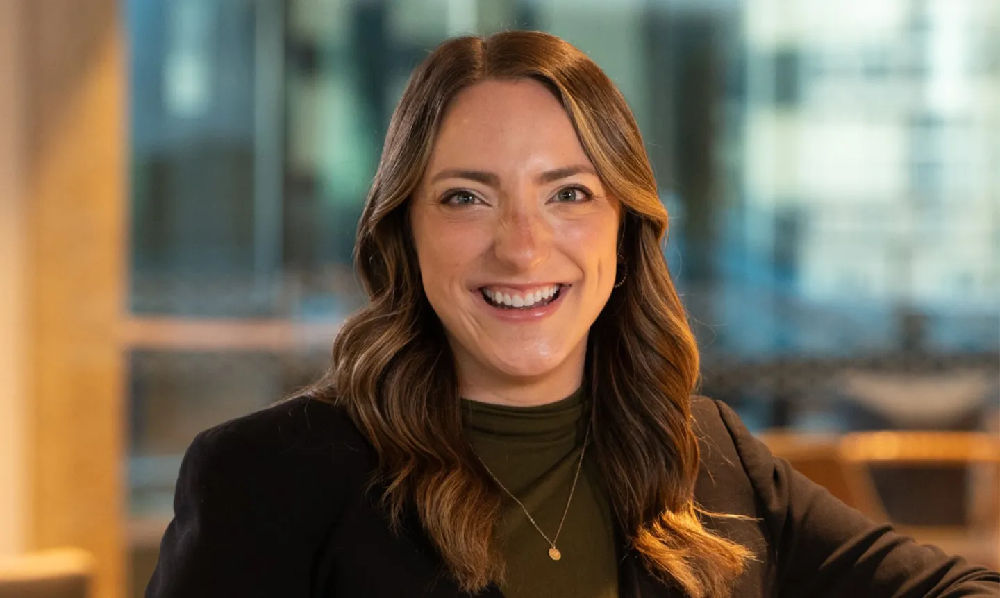 A portrait of Maura Bremer Peterson. She is smiling and wearing an olive green turtleneck, brown blazer, and a gold necklace. Her hair is down.