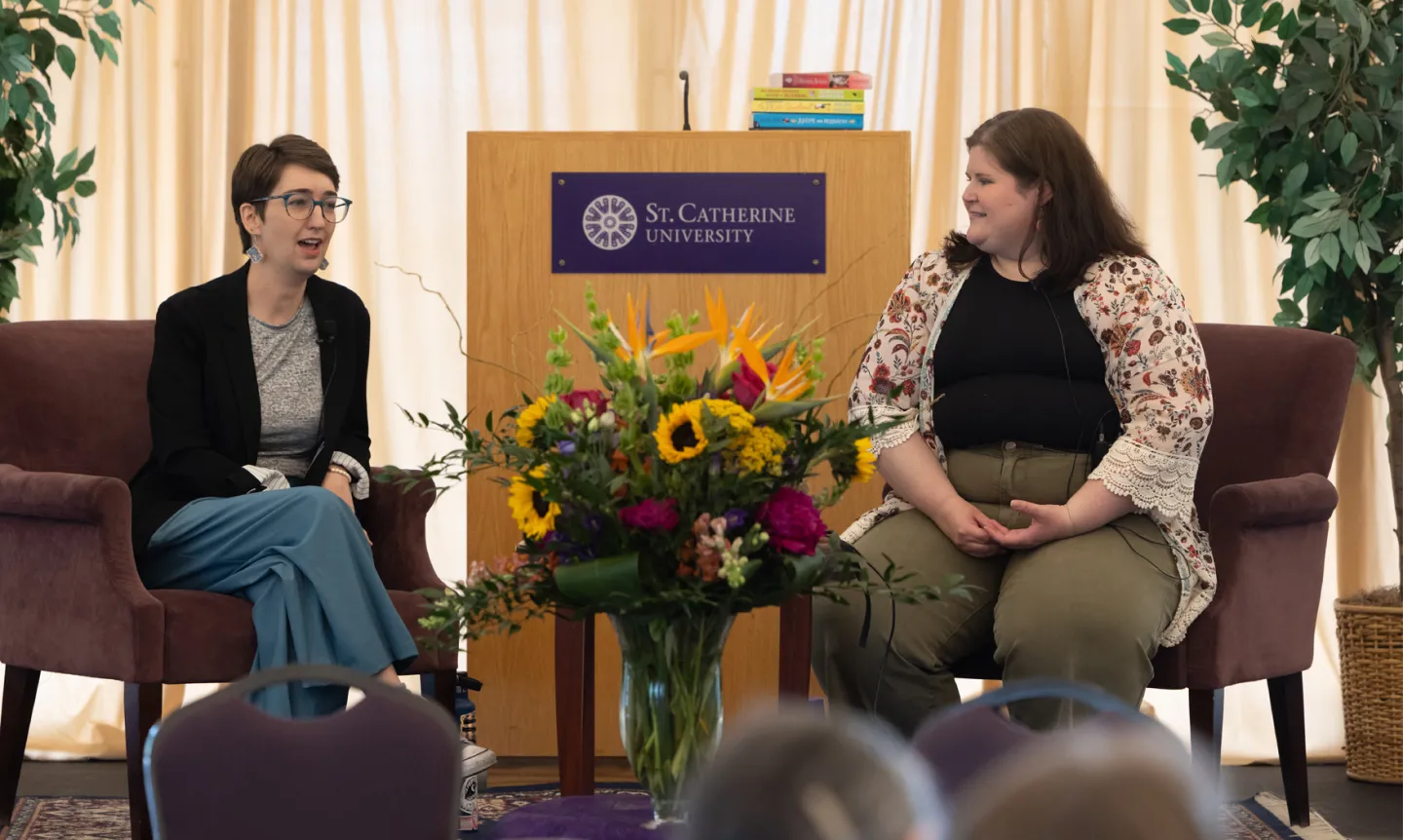 Two women sit in armchairs talking. The woman on the left has short brown hair, glasses, a black blazer over a gray shirt, and turquoise pants. The woman on the right has medium-length brown hair, a floral cardigan over a black shirt, and olive green pants. Between them is a vase of flowers and a podium.