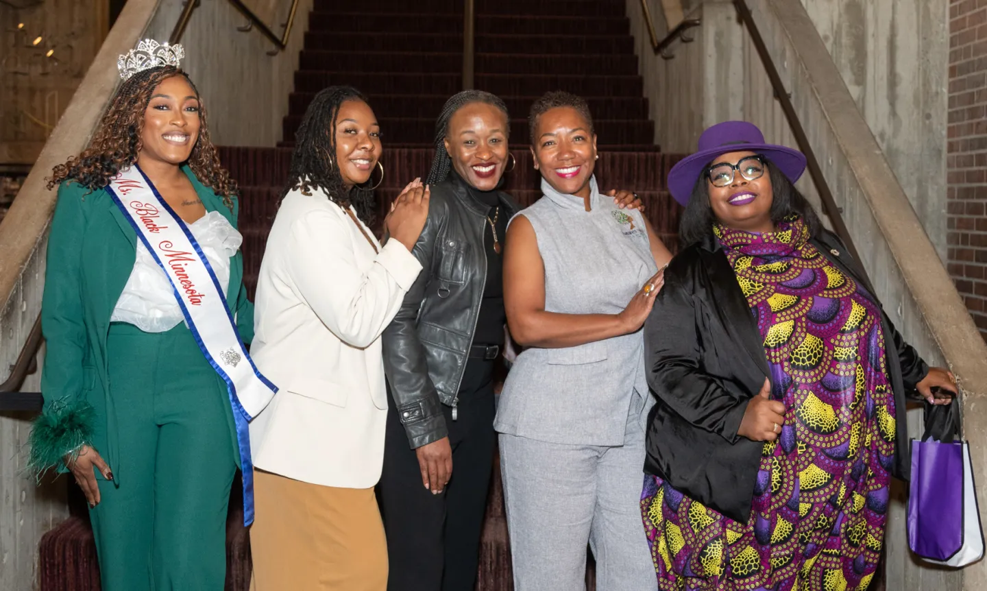 A group of five women standing together in front of stairs inside a building.