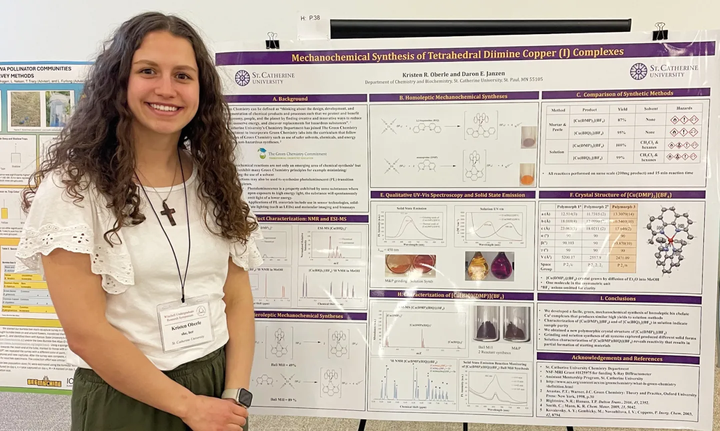 Photo of a Kristen Oberle standing in front of a large poster displaying scientific research and data. Her hair is long and curly, she is smiling, and she is wearing a white blouse with green pants, a cross necklace, and a nametag that reads "Winchell Undergraduate Research Symposium. Kristen Oberle, she, her, St. Catherine University."