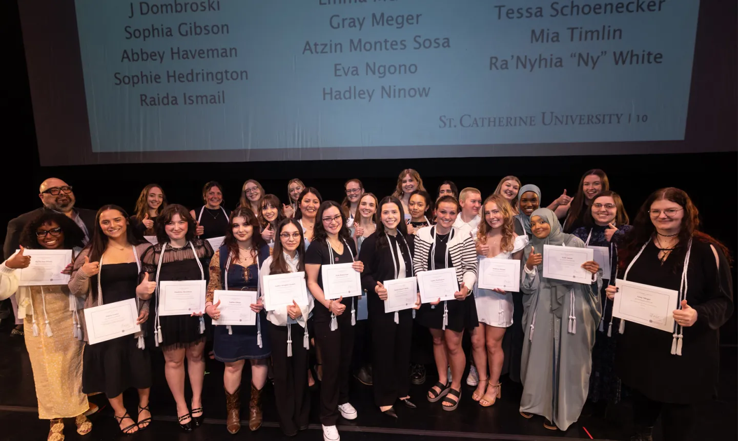 The Antonian Scholars stand on stage, smiling and holding up paper certificates.