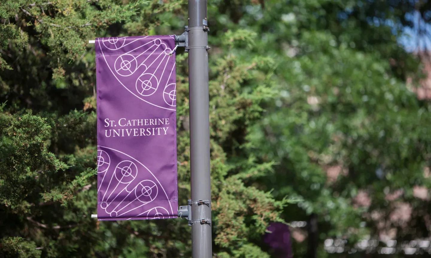 A purple flag reading "St. Catherine University" hangs on a pole in front of a background of trees.