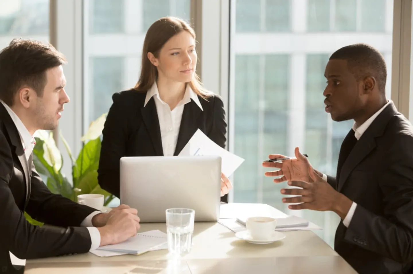 A trio of two businessmen and one businesswoman intently discuss a project in a high-rise business office.