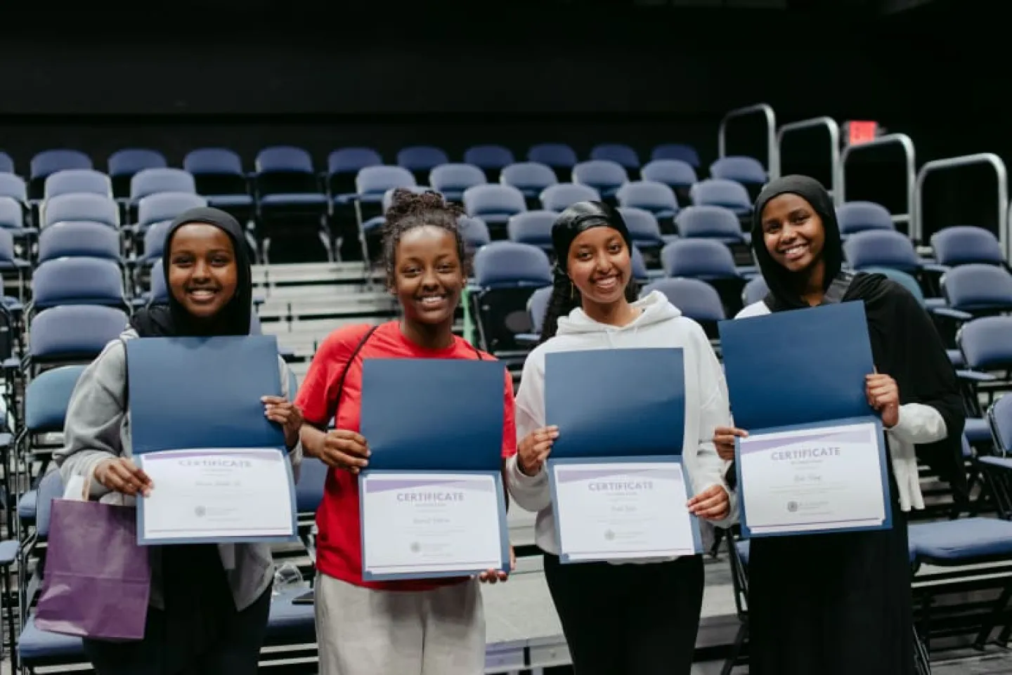 Four students standing in an auditorium holding certificates and smiling at the camera.