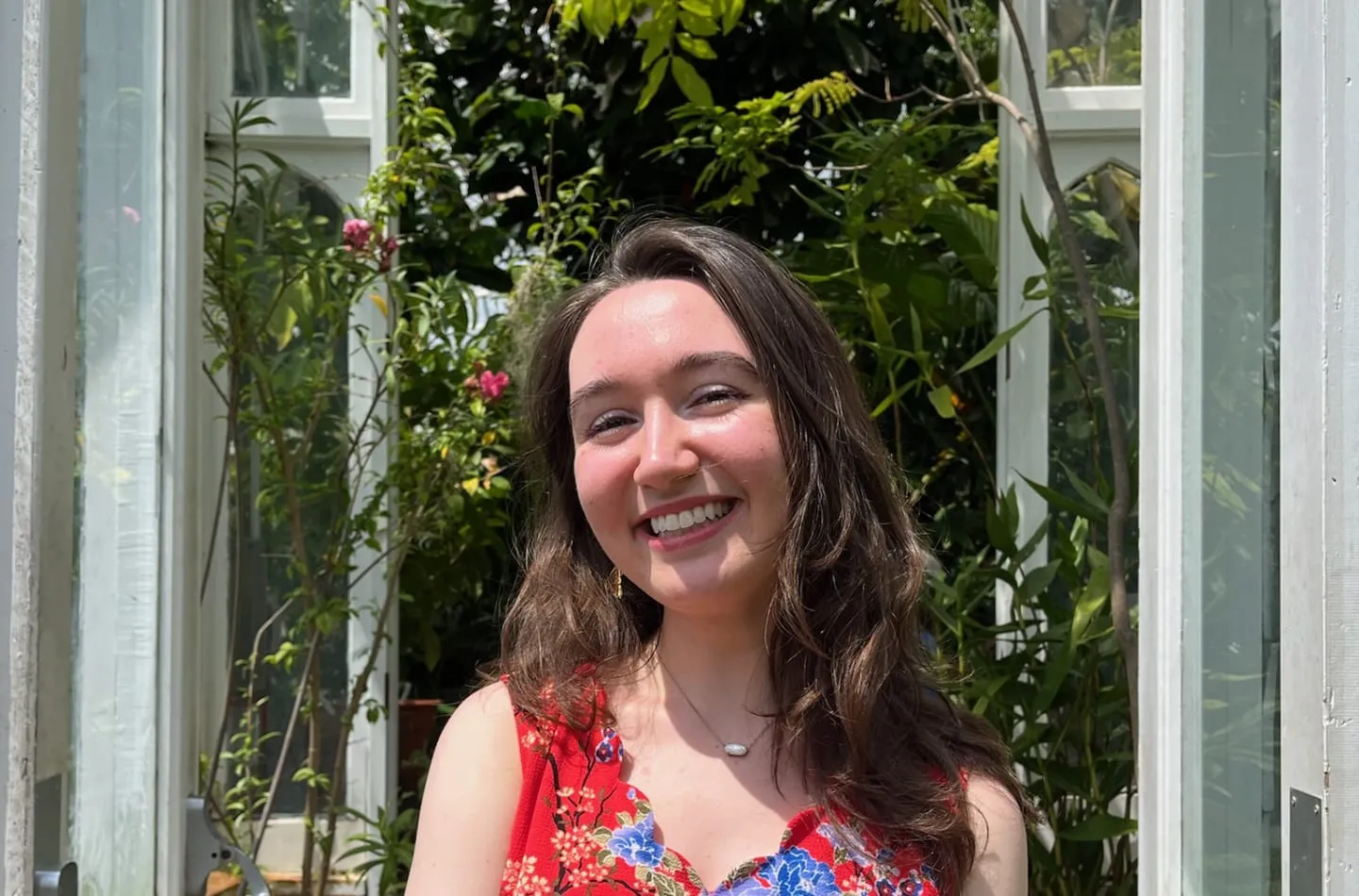 Smiling young woman with long brown hair wearing a red floral dress, standing in front of open glass doors with lush greenery and flowers in the background.