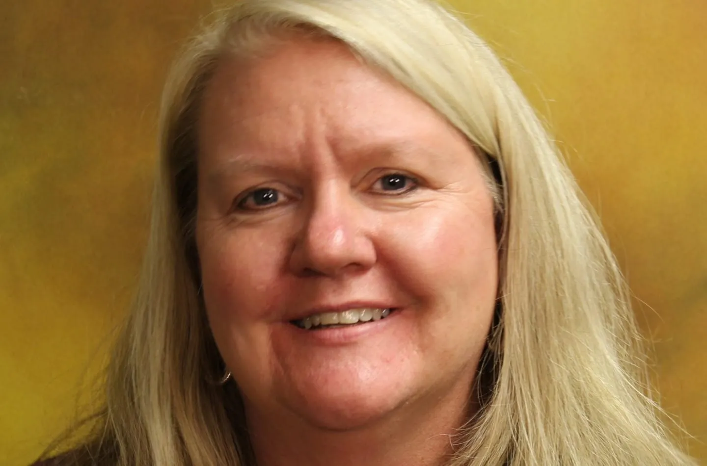 Portrait of Kim Devlin smiling with long blonde hair, against a yellow and brown studio background.