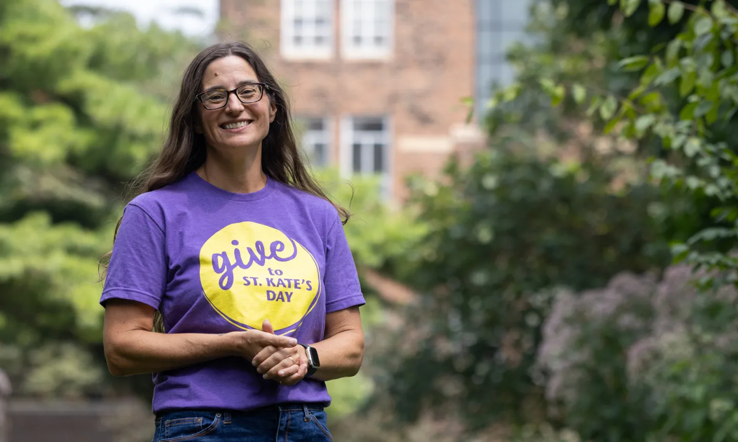 A smiling woman with glasses wears a purple Give to St. Kate's Day T-shirt.