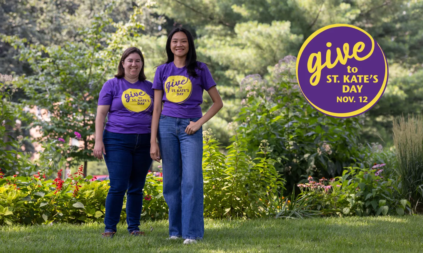 Two people wearing purple T-shirts and jeans stand in a green, leafy setting. A logo reads "Give to St. Kate's Day Nov. 12"