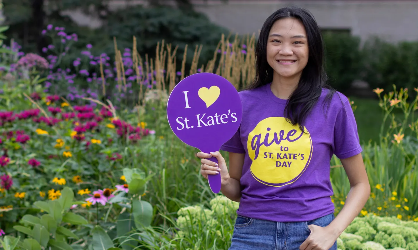 A smiling student wears a purple "Give to St. Kate's Day" shirt and holds up a small sign that reads "I Heart St. Kate's"