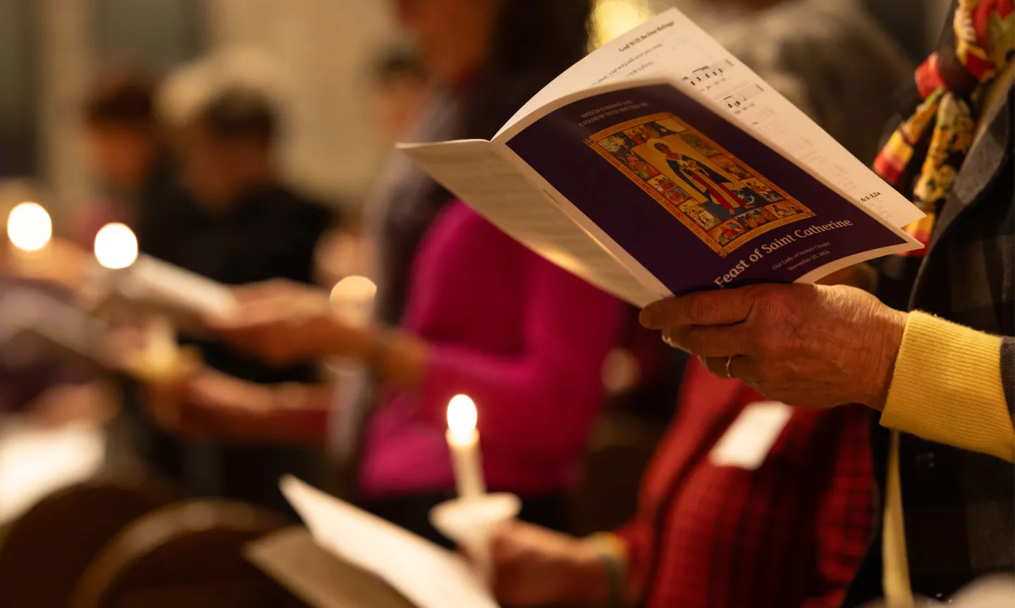 At the Feast of Saint Catherine prayer service in the Chapel, hands hold a prayer book. Lit candles can be seen in the blurred background.