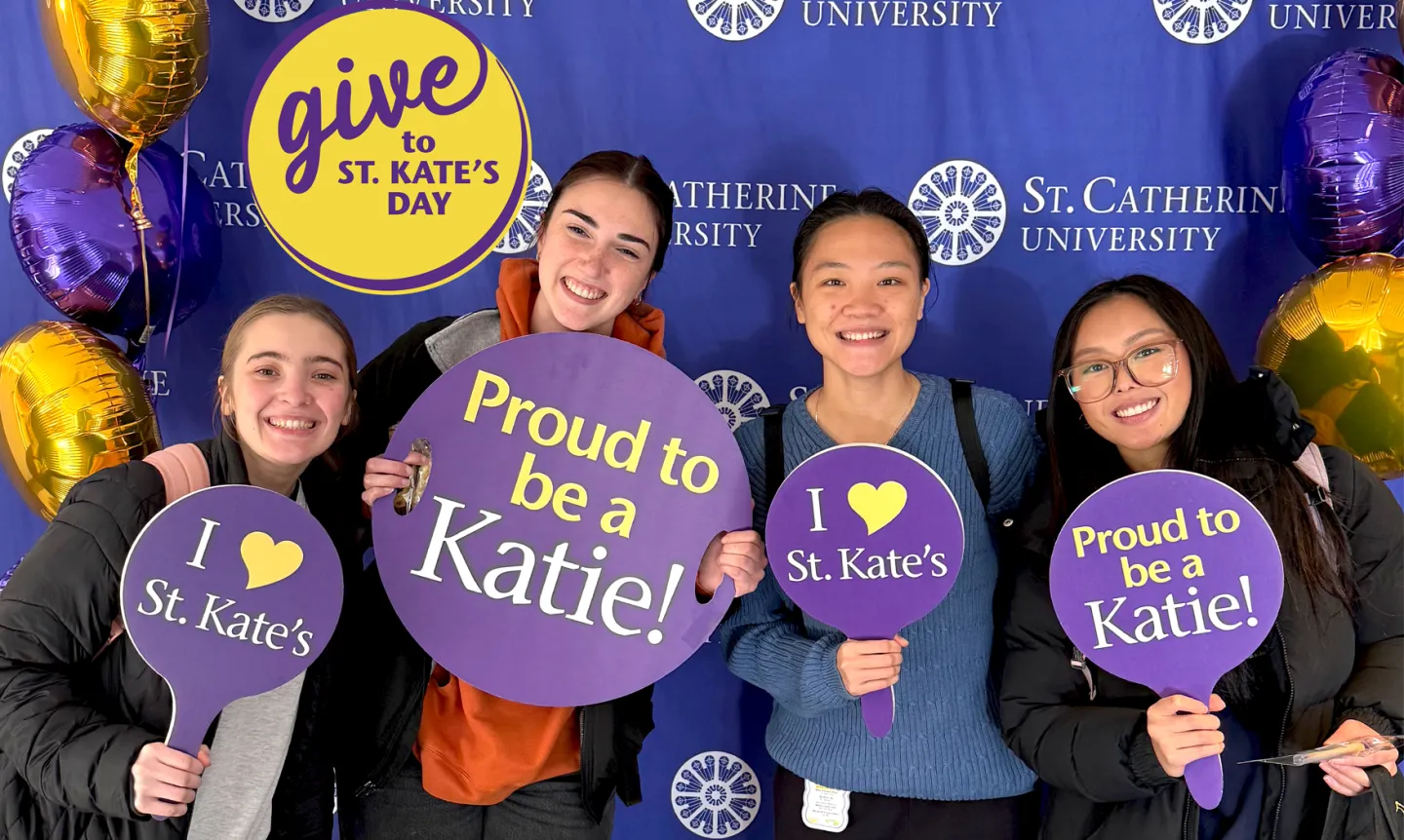 Four students pose with purple signs that say "Proud to be a Katie!" and "I heart St. Kate's"