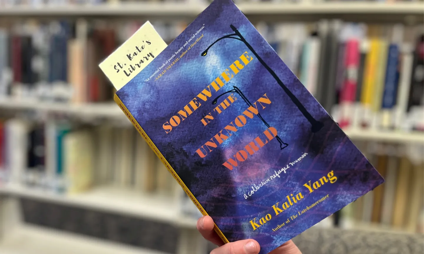 A hand holds a copy of "Somewhere in the Unknown World," with St. Kate's library shelves in background