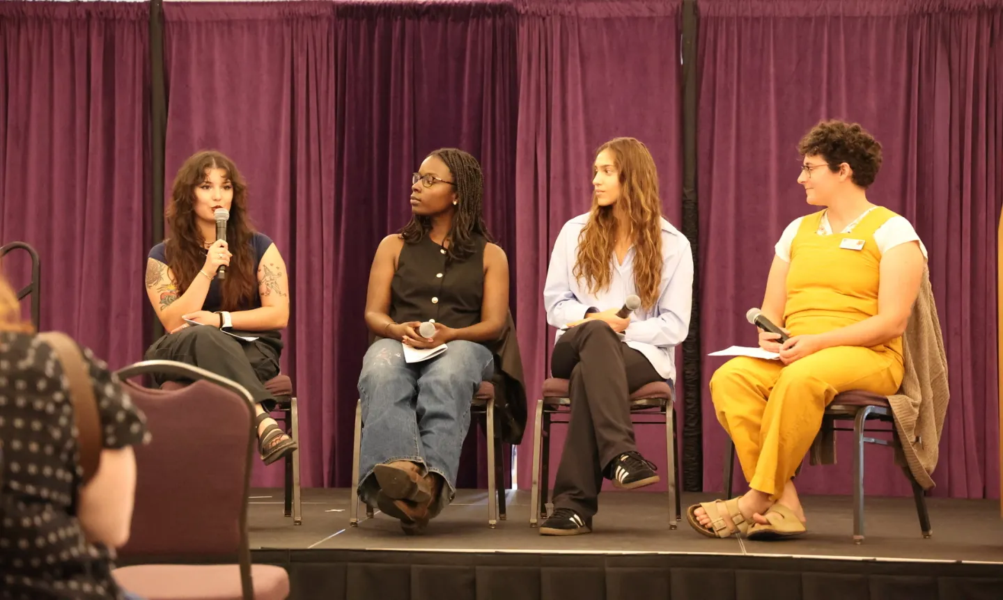 Four members of a panel sit on a stage; one of them speaks into a microphone.