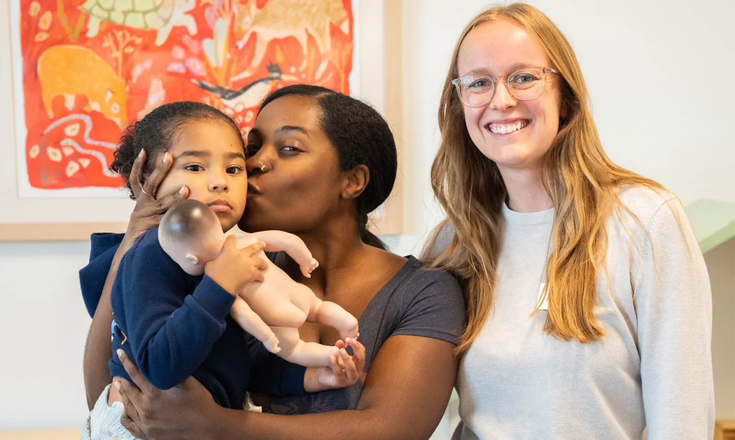 Two women stand and smile; one of them holds a child in her arms.