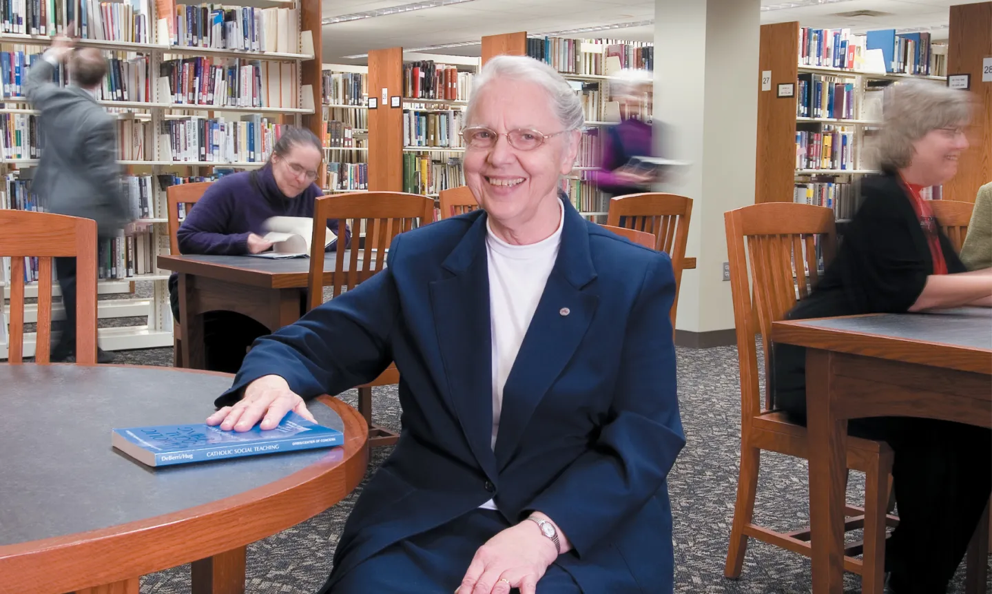 Sister Amata Miller sits at a library table with books and people behind her.