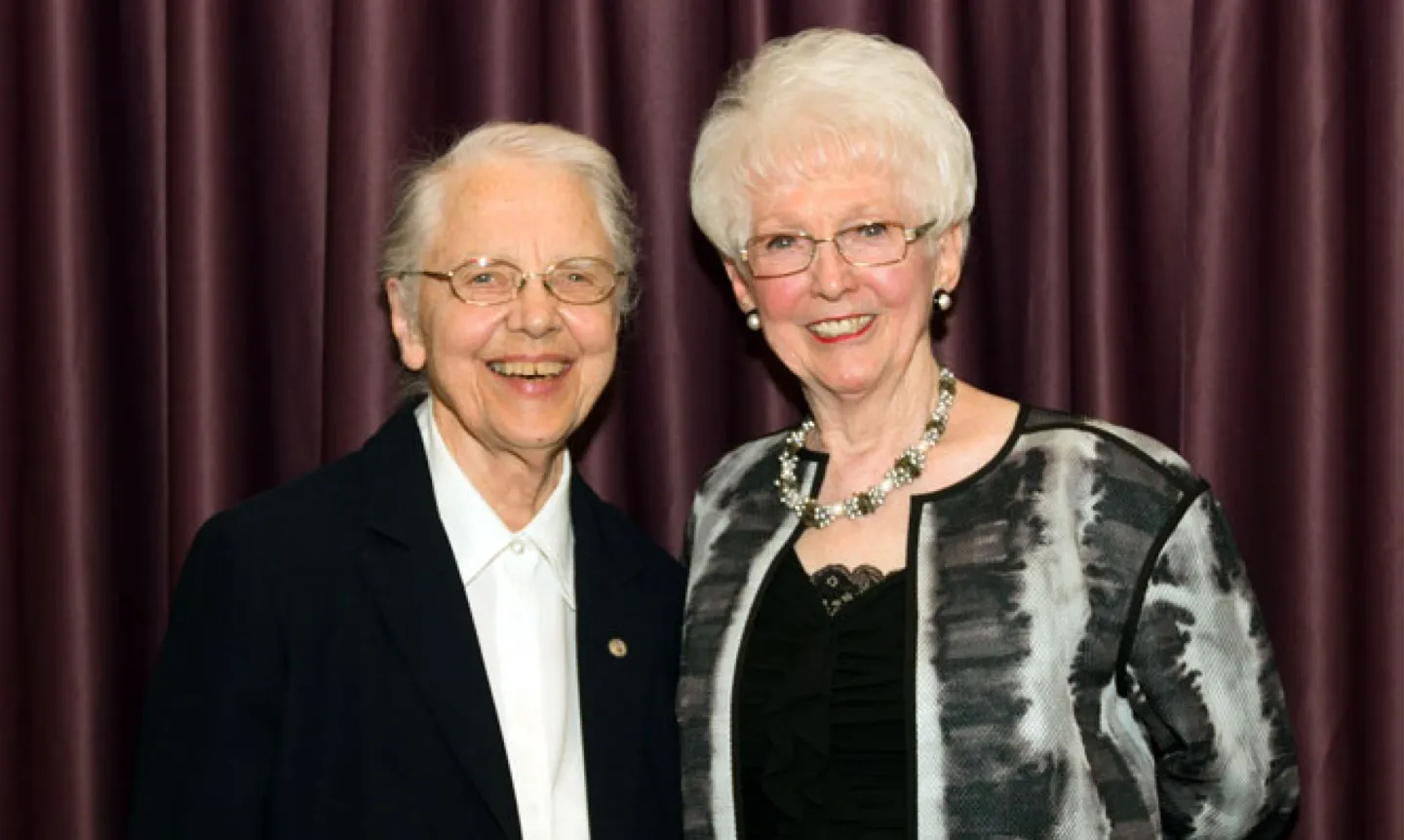 Sister Amata Miller and Patrician O'Connor Myser pose together in front of a maroon curtain.