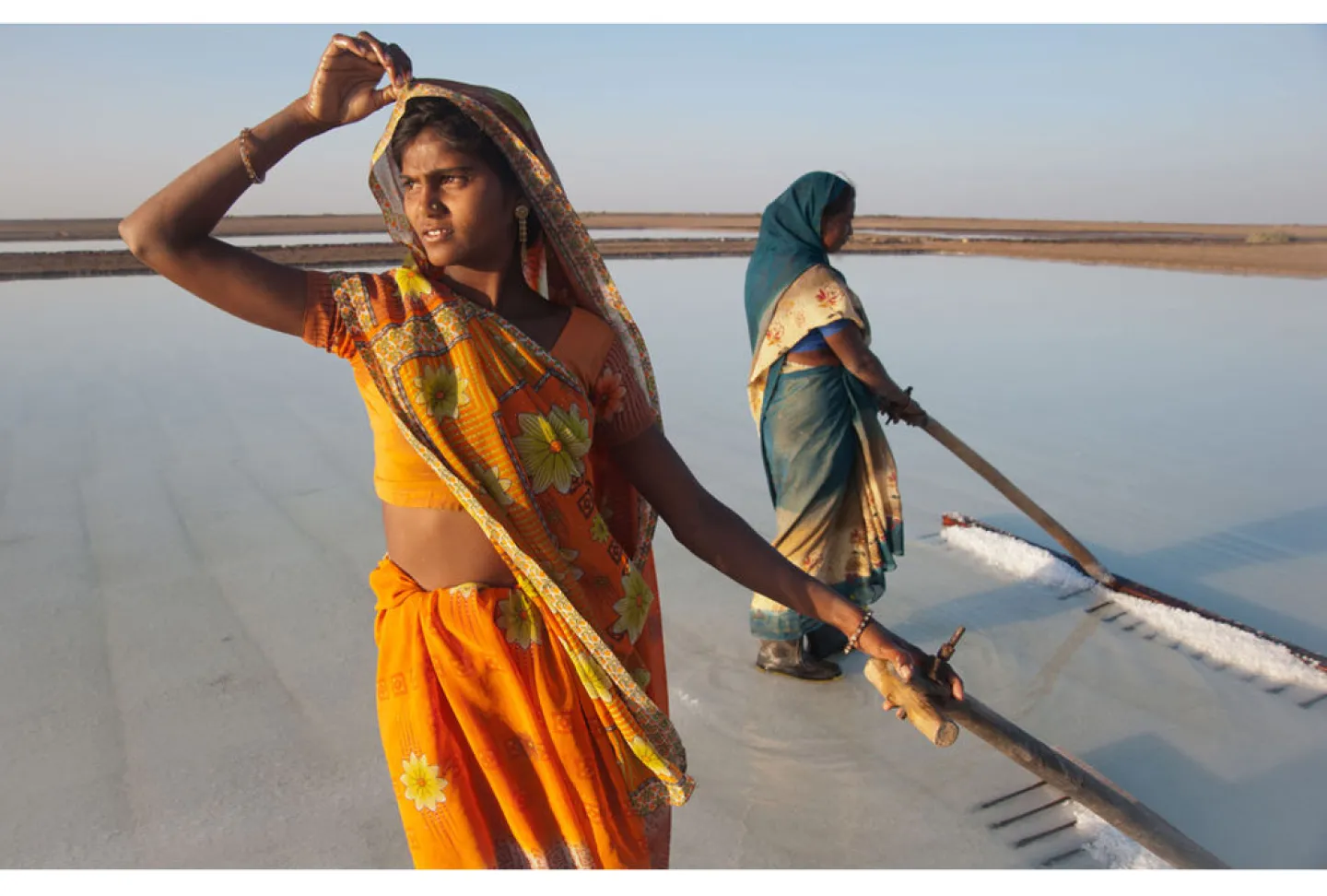 Photo title is "Ripple Effect Image" - shows two (possibly Indian) women in saris standing on sand in ankle-deep water, and using tools that look like large rakes