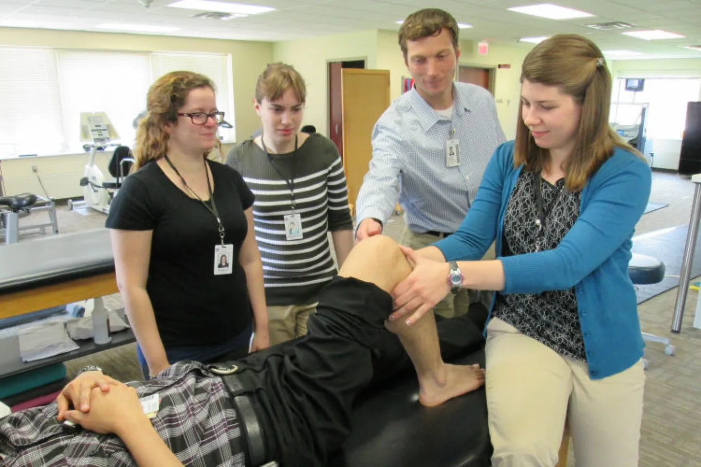 Photo of five people. One (probably a student playing the part of a patient) is lying down, with right leg flexed and pants leg pushed up to expose that leg from the knee down. Two people (a student and a faculty member?) are assisting the patient, and two others observe the demonstration.