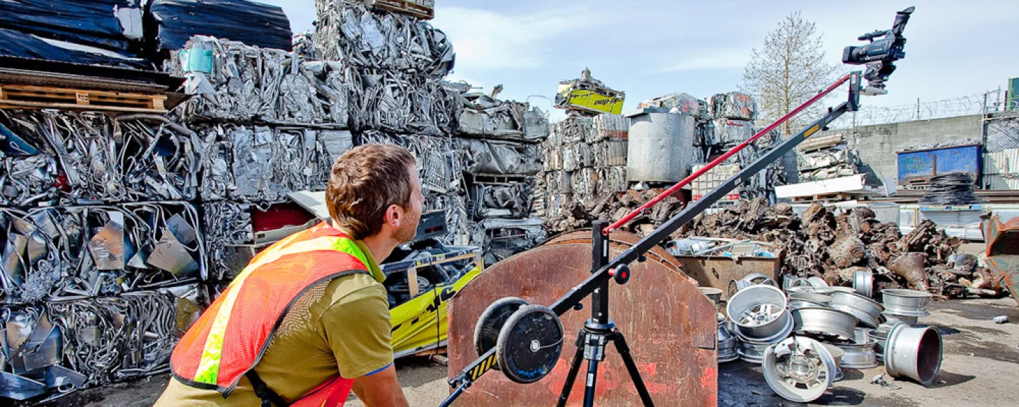 Photo of a man operating a movie camera in an industrial area with stacked bales of crushed metal (auto parts being prepared for recycling?)